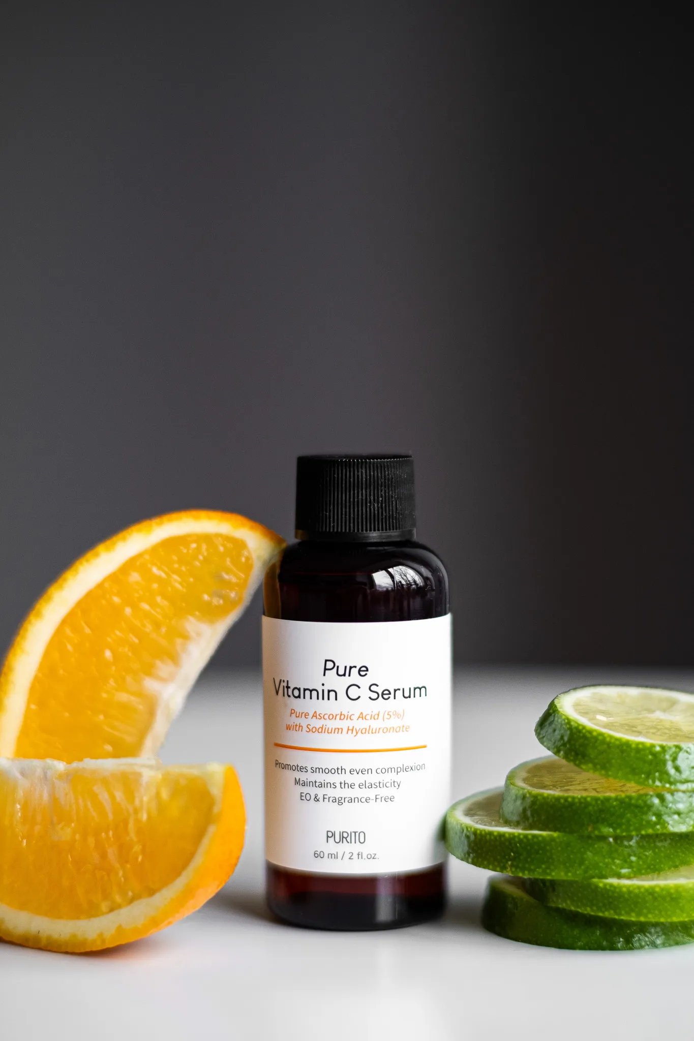 Предметна фотографія пляшечки сироватки Pure Vitamin C Serum на білому фоні з апельсином і лаймом.