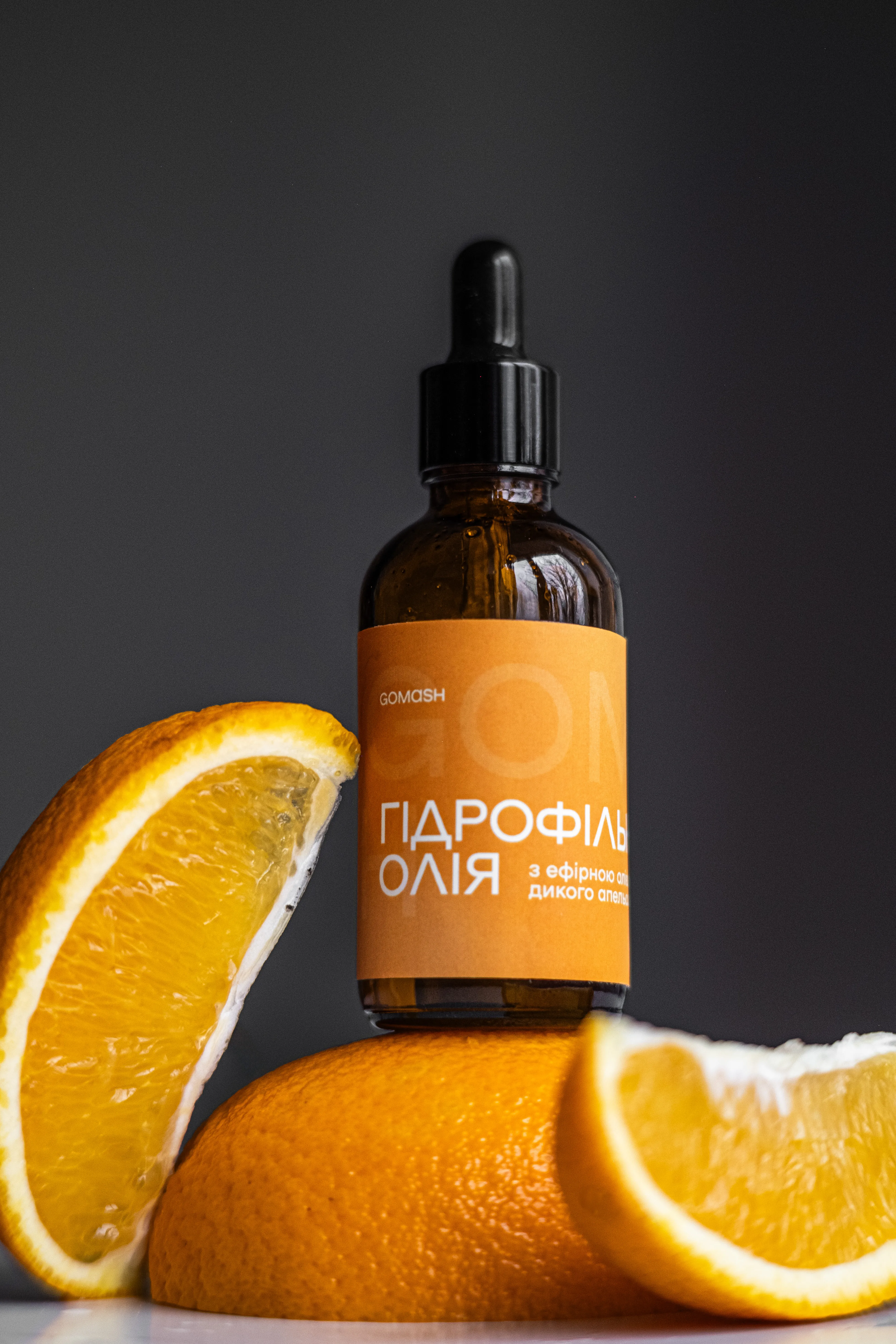 Предметна фотографія гідрофільної олії з ефірною олією дикого апельсина у скляній упаковці на тлі апельсинів.