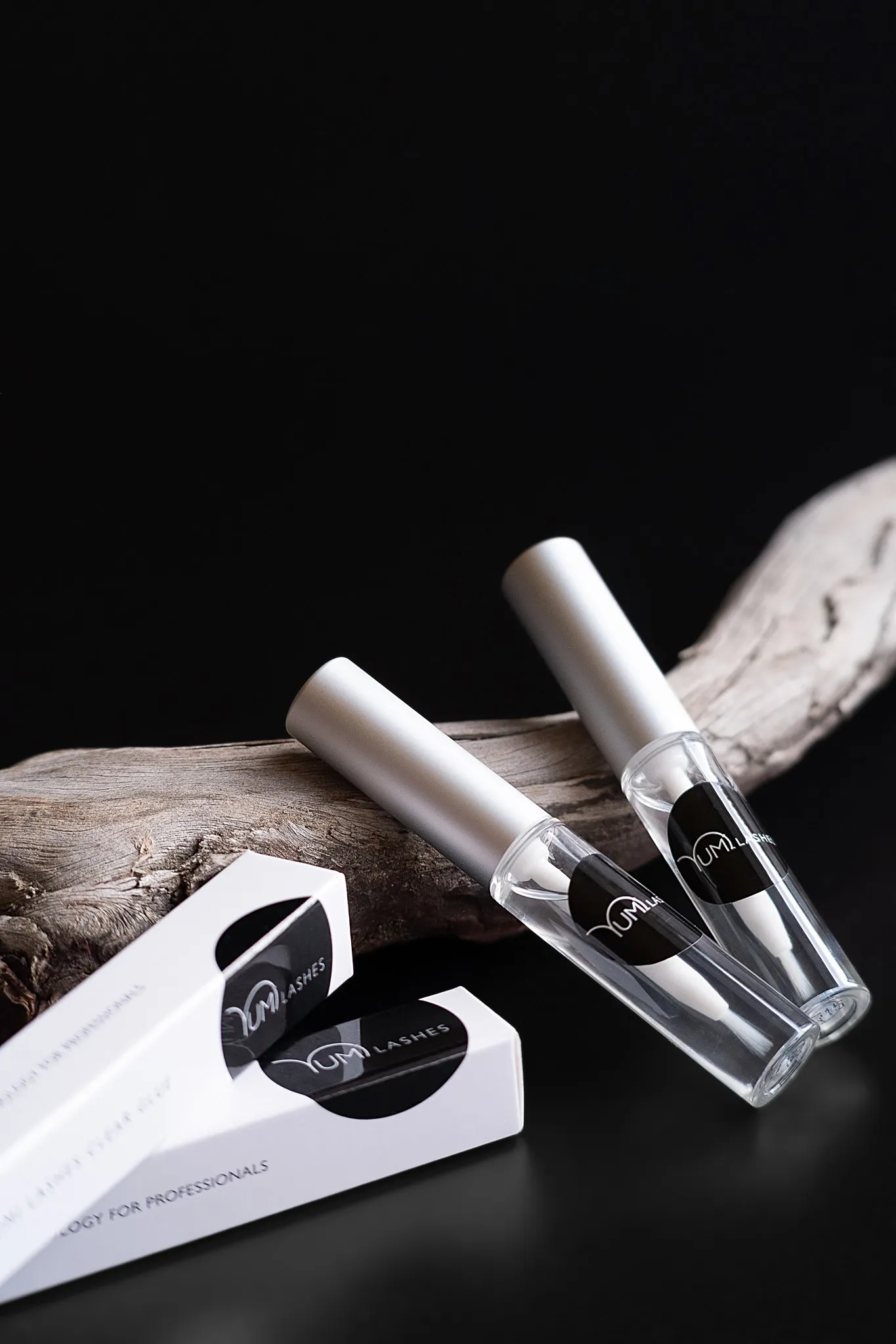 Предметна фотографія двох тюбиків косметики для вій YUM Lashes на чорному фоні з гілкою сухого дерева.