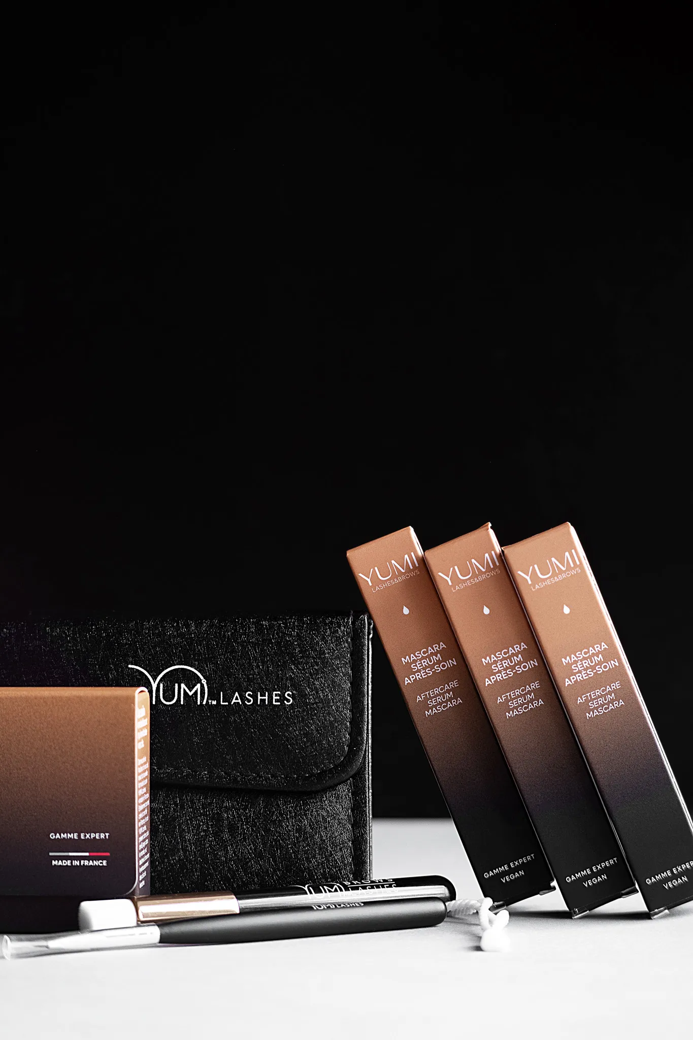 Предметна фотографія косметики YUMI Lashes: туш, сироватка та аксесуари для вій і брів на білому тлі.