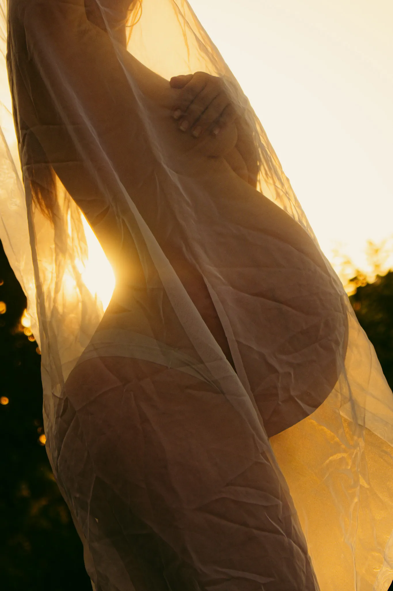 Вагітна жінка під прозорою тканиною на заході сонця – сімейна та maternity фотосесія на природі.