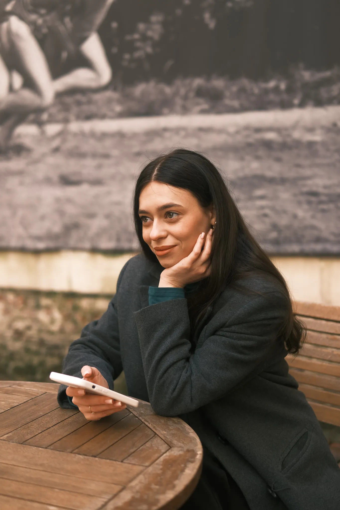 Жінка в пальті тримає смартфон, сидячи за столом на вулиці. Індивідуальна зйомка на природі.