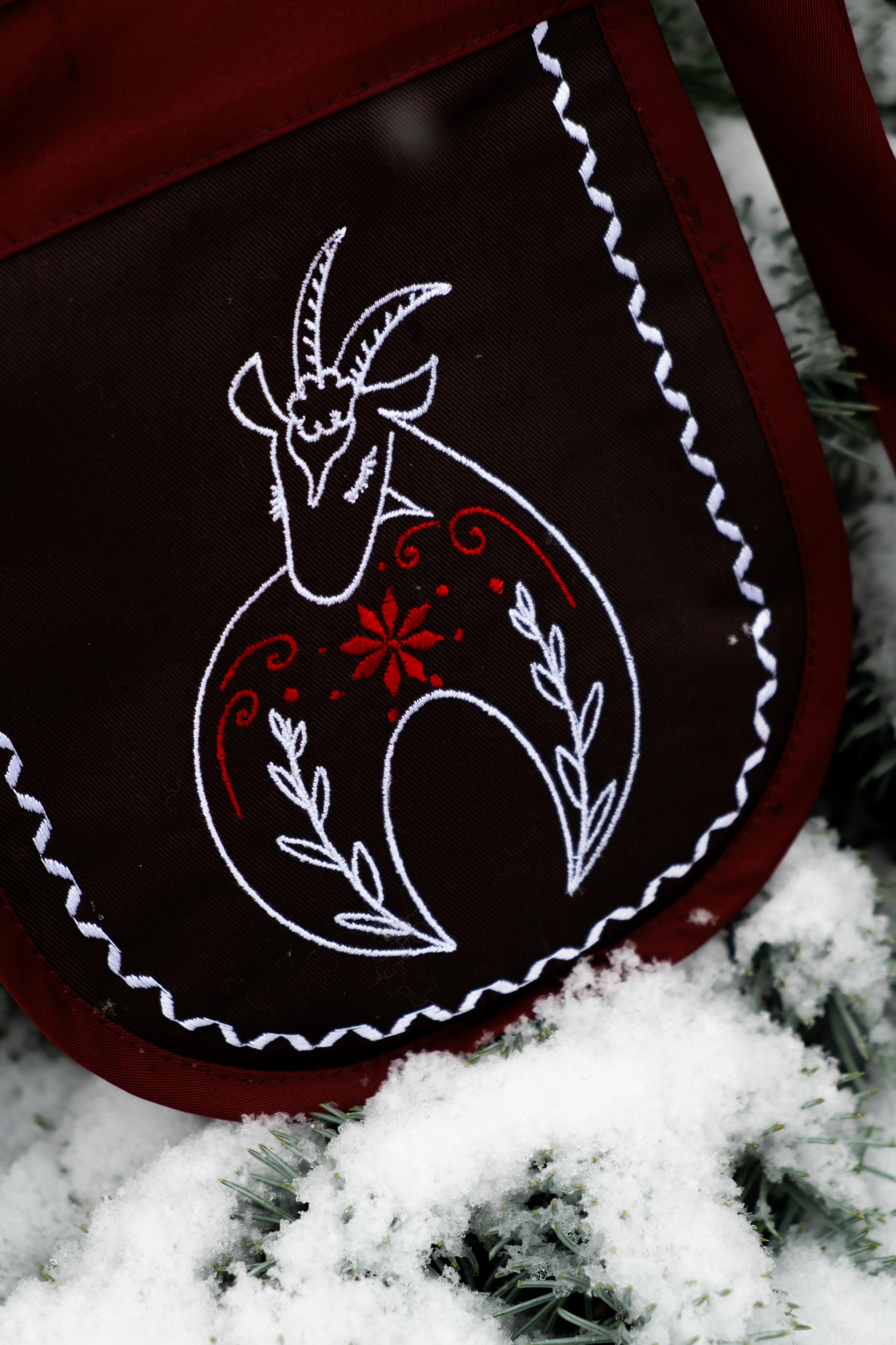 Предметна фотографія коричневої сумки з вишивкою кози, біло-червоні деталі, зимовий фон зі снігом.