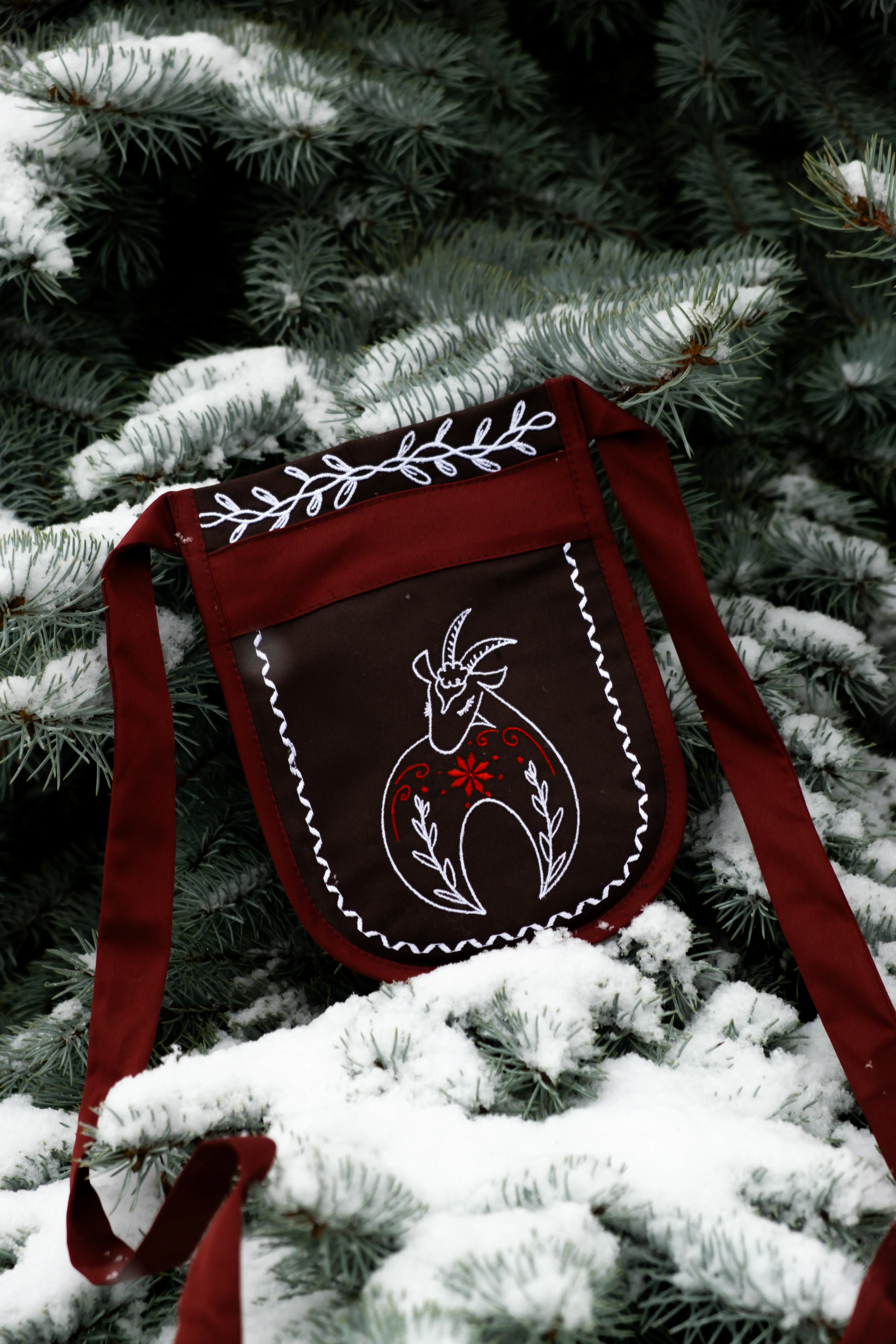 Предметна фотографія: бордова сумка з вишивкою козла на фоні ялинкових гілок із снігом.