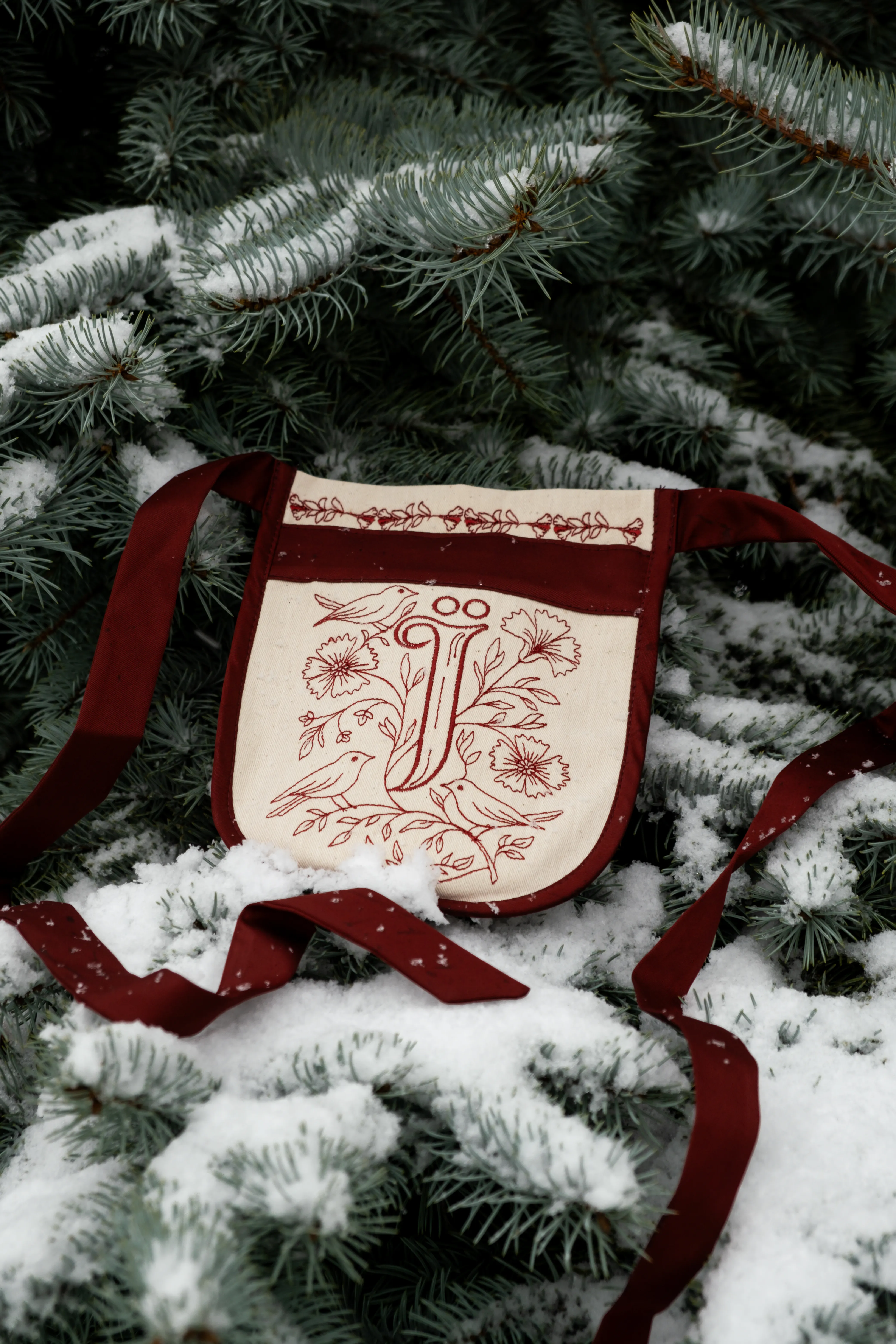 Предметна фотографія: вишитий льняний фартух з鳥ми на фоні засніжених ялинових гілок.