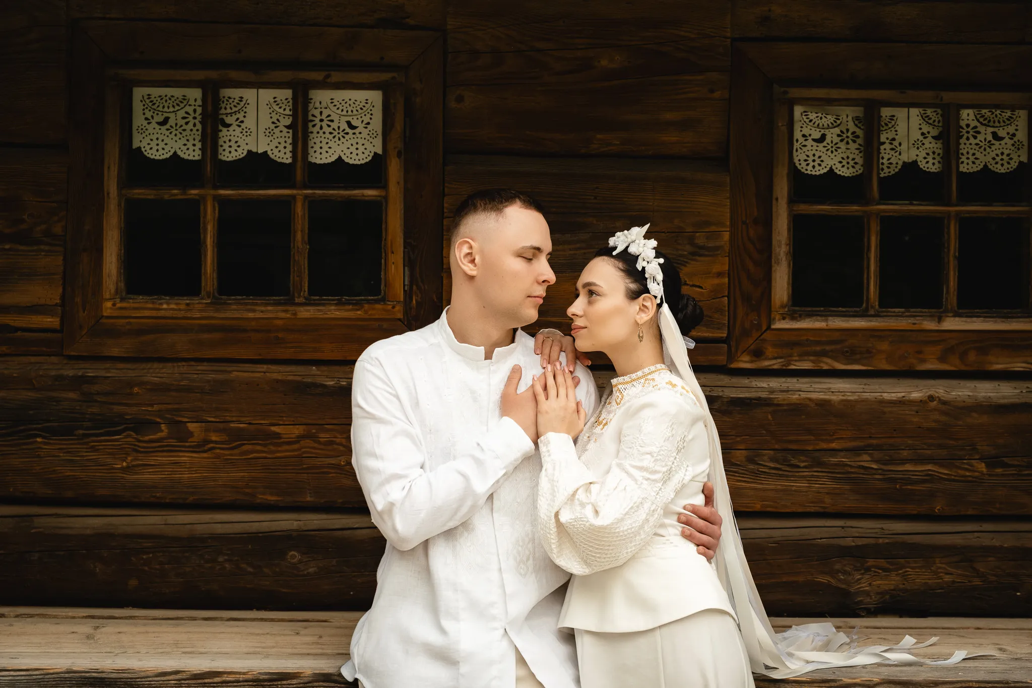 Молодята фото на фоні дерев'яного будинку у традиційному українському вбранні, весільний портрет закоханої пари.