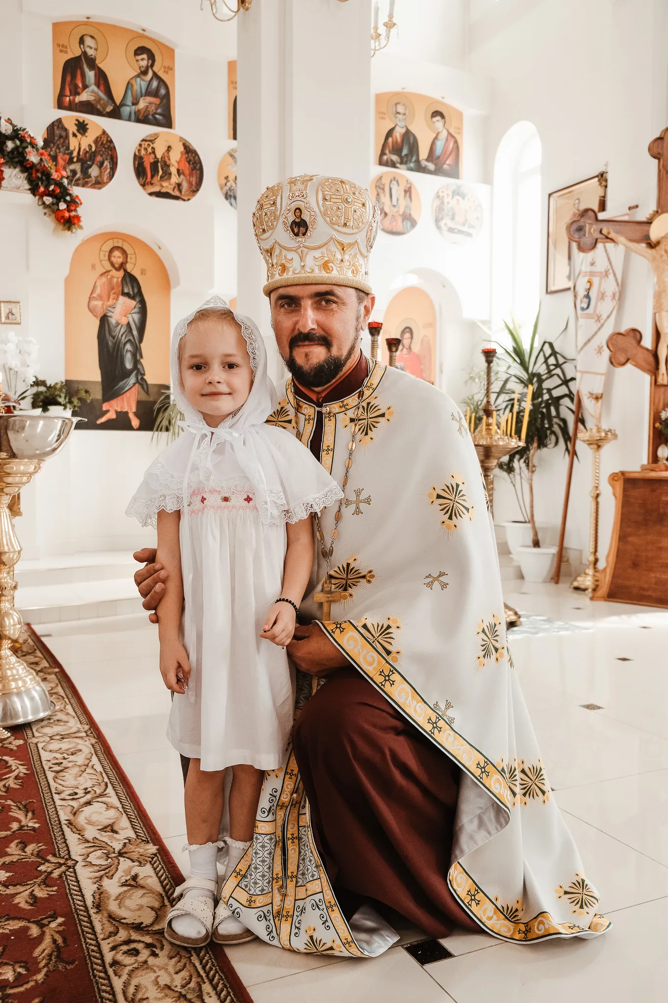 Священник у храмі на Таїнстві Хрещення з дівчинкою у білій сорочці та хустці, релігійне свято, церковне фото.