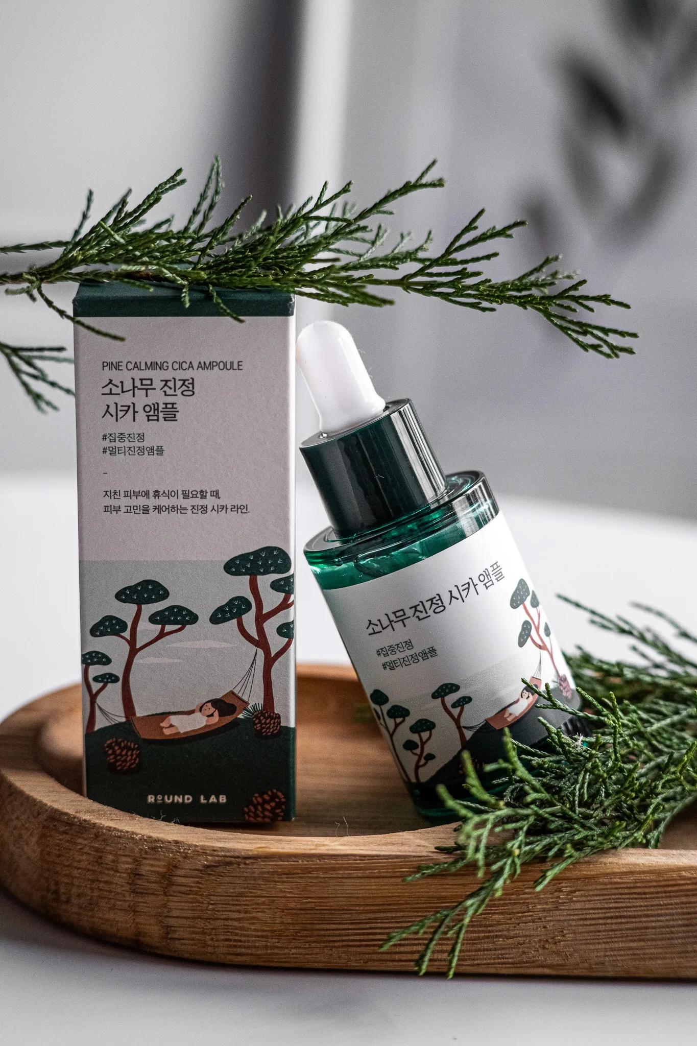 Предметна фотографія: косметична сироватка Pine Calming Cica Ampoule на дерев'яній підставці з гілками ялини.